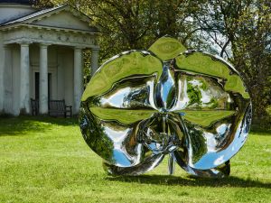 A large sailver abstract sculpture sits in a park like space