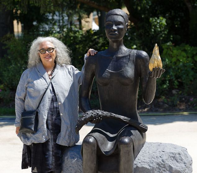 A woman with long gray hair poses next to a large statue of a woman holding a flame