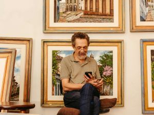 A man sits looking at a phone in front of a wall of framed artworks