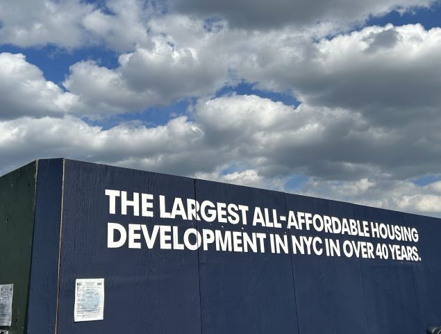 Construction barrier with bold white text reading "The largest all-affordable housing development in NYC in over 40 years" against a backdrop of a partly cloudy sky.