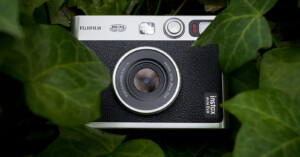 A Fujifilm Instax Mini Evo camera with a retro design is nestled amongst green leaves. The camera’s lens and buttons are visible, surrounded by the foliage.