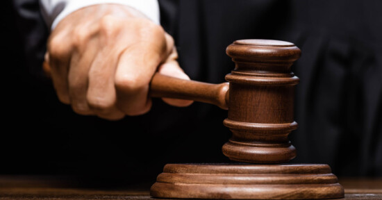 A close-up view of a person's hand holding and striking a wooden gavel on a sounding block, symbolizing a decision or judgment being made in a courtroom setting. The background is partially visible and appears to be a dark judicial robe.