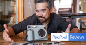 A person is sitting at a wooden table, smiling, and holding a Fujifilm Instax Wide 300 instant camera. Several instant photos are scattered on the table in front of them. The image includes the PetaPixel Reviews logo. The background shows a brightly lit interior.