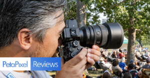 A person with short gray hair is closely holding and looking through a professional camera. The scene is outdoors with trees and a crowd in the background. The image has banners with the text "PetaPixel" and "Reviews.