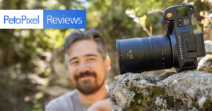 A camera with a large lens is perched on a rock outdoors. In the background, a person with a beard and mustache is slightly out of focus, smiling and looking at the camera. The text "PetaPixel Reviews" is displayed in the upper left corner.