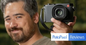 A person holding a camera close to the lens, slightly out of focus. The background is blurred greenery. There's a logo for PetaPixel Reviews in the lower right corner.