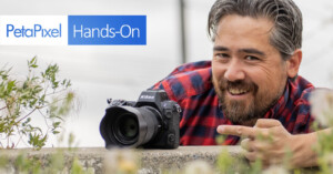 A person with a beard, wearing a red and black checkered shirt, leans over a wall and points to a Nikon camera placed on it. The text "PetaPixel Hands-On" is visible in the top left corner of the image.