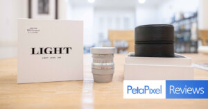 A photography lens on a table, accompanied by its packaging box and a storage case. The box features "LIGHT" branding and a small text, "Petapixel Reviews," is overlaid on the bottom right. The background is an out-of-focus room.