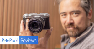 A person holding a vintage-style camera close to the lens, standing against a blurred wooden background. The image features the text "PetaPixel Reviews" in the bottom left corner.