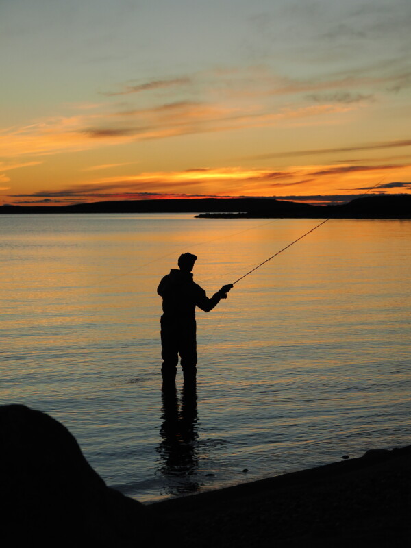 Silhouette of a person fishing in calm waters during a sunset. The sky is painted with warm shades of orange and yellow, reflecting on the water. The scene is tranquil and serene, capturing a peaceful moment by the shoreline.