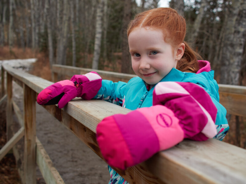 A young girl with red hair wearing a teal jacket and pink mittens leans on a wooden railing along a forest path, smiling. The background shows bare trees and the remains of melting snow.