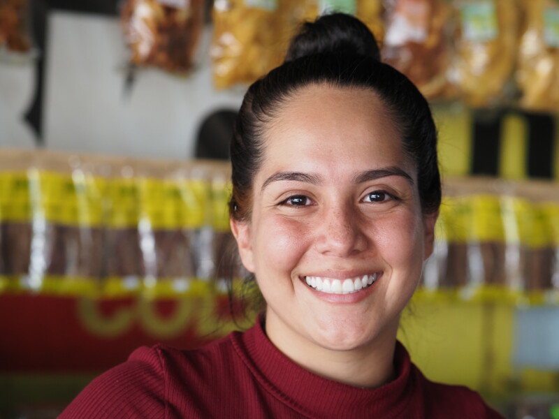 A person with long dark hair in a bun smiles warmly at the camera. They are wearing a maroon top. The background shows packaged products on a display, slightly out of focus.