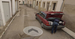 A person leans into the trunk of a parked red station wagon on a narrow, empty street lined with beige buildings. The road has a weathered appearance, and the area looks quiet and residential.