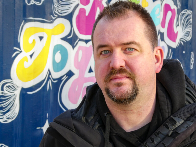 A person with short hair and a beard is standing in front of a colorful mural with stylized lettering. They are wearing a dark jacket and looking directly at the camera. The mural features vibrant colors and intricate designs.