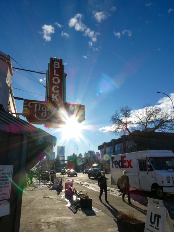 Street scene with people walking past a FedEx truck. A vintage-style neon sign reads "BLOCK" against a bright sun and partly cloudy sky. The urban setting includes a mix of building styles and bare trees.