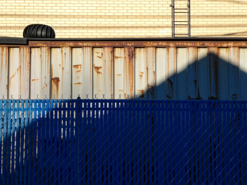 A rusty metal wall casts a shadow in the shape of a triangle. A blue chain-link fence stands in front. In the background, a yellow brick building with a rooftop ladder and a black vent is visible.