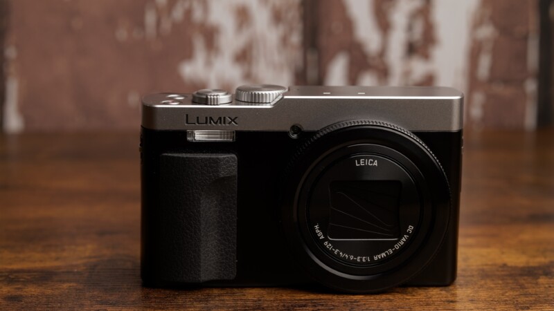 A black and silver Lumix camera with a Leica lens sits on a wooden surface. The background is a blurred mix of white and brown tones.