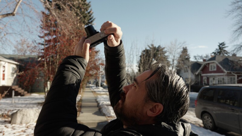 A man in a black jacket holds a camera pointed towards the sky on a sunny winter day. He stands on a snow-dusted sidewalk lined with trees and houses.