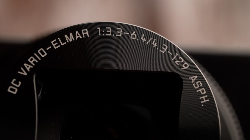 Close-up of a camera lens with the text "DC VARIO-ELMAR 1:3.3-6.4/4.3 12.9 ASPH." engraved on the edge. The lens surface has concentric circular patterns, creating a textured appearance. The background is softly blurred.