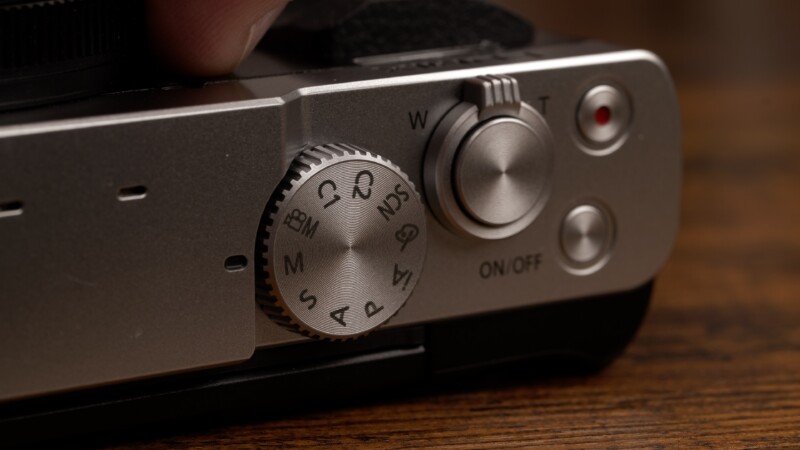 Close-up of a camera's mode dial and power switch. The dial shows options like "M," "A," and "C" for different shooting modes. Next to it is a power switch labeled "ON/OFF" and a zoom button marked "W" and "T". The background is wooden.