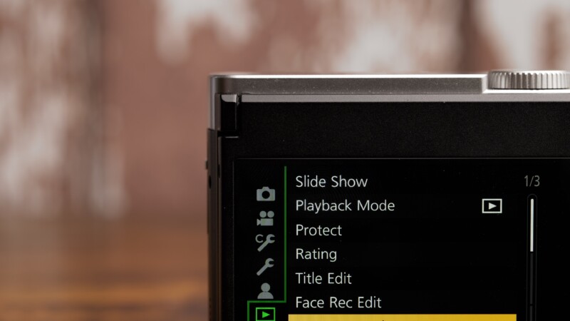 Close-up of a digital camera screen displaying menu options, including Slide Show, Playback Mode, Protect, Rating, Title Edit, and Face Rec Edit. The background is blurred with a warm brown tone.