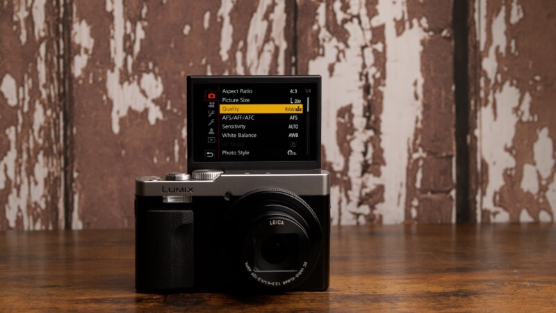 A Panasonic Lumix camera sits on a wooden surface in front of a rustic, weathered wooden wall. The camera's LCD screen is flipped up, displaying its menu interface with various settings options.