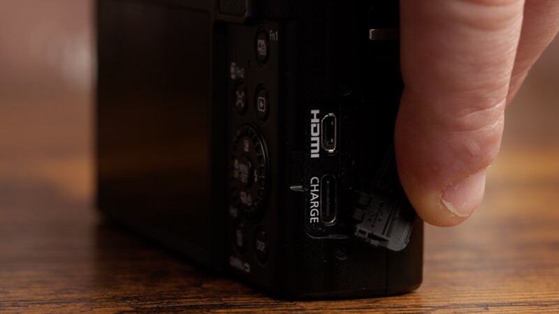 A person plugs a cable into the HDMI and charge port of a black camera. The camera is positioned on a wooden surface, and the person's thumb is visible holding the plug.