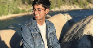 A person wearing glasses and a denim jacket is smiling while sitting on a large rock by a river. The background features trees and boulders in a sunny setting.