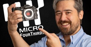 A man smiles and points at a camera he is holding. The camera has no lens attached. Behind him, a logo reads "Micro Four Thirds" on a dark background.