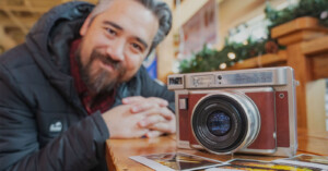 A person leans forward with a smile beside a large vintage camera on a wooden table. Several instant photos are scattered on the table. The background shows part of a warmly lit interior with festive decorations.