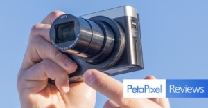 A person holding a compact camera with a zoom lens extended. The camera lens has "Leica" branding. The image has a blue sky background and features a "PetaPixel Reviews" graphic in the bottom right corner.