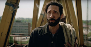 A man with a beard and dark hair stands outdoors, holding a coiled rope over his shoulder. He is wearing a dark jacket. A construction site with metal beams and a distant building can be seen in the background.