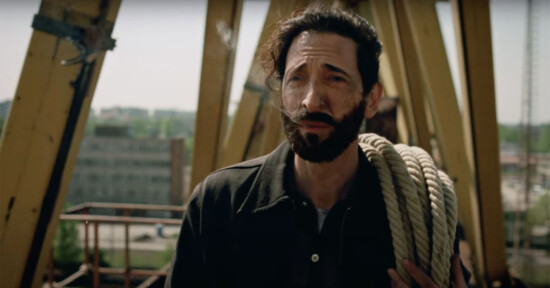 A man with a beard and dark hair stands outdoors, holding a coiled rope over his shoulder. He is wearing a dark jacket. A construction site with metal beams and a distant building can be seen in the background.