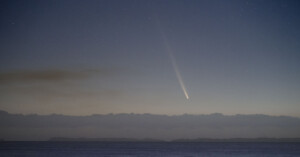 A comet with a long, bright tail streaks across a starry night sky above the horizon, casting a serene glow over the calm ocean below. Dark silhouettes of distant landmasses are visible at the horizon line.