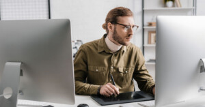 A person with a beard and glasses works at a desk using a drawing tablet, surrounded by two large computer monitors. They are wearing a green shirt and appear focused on their task in a bright office space.