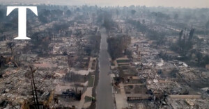 Aerial view of a devastated neighborhood after a wildfire. Charred remains of homes and trees line both sides of an empty street. Smoke lingers in the air, and the scene appears desolate and heavily damaged.