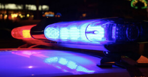 Close-up of a police car light bar at night, with blue and red lights illuminated, reflecting on the vehicle's surface. Blurred lights are visible in the background.