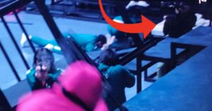 Several people in green uniforms are crawling on the floor under a structure, while a person in a pink outfit with a mask observes them. A large red arrow is pointing towards one of the individuals in green. The scene is dimly lit.