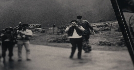 A black and white photo shows a group of people outdoors. Two individuals in focus are looking at cameras, while another person walks away in the background. The scene appears to be candid, with a blurred landscape behind them.