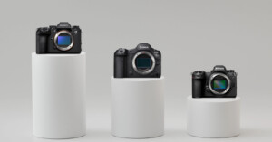 Three digital cameras are displayed on white cylindrical pedestals. From left to right: a Sony camera, a Canon camera, and a Nikon camera, each with a visible lens mount and positioned on varying pedestal heights against a neutral background.