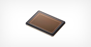 A close-up image of a rectangular camera sensor chip with a metallic frame and numerous gold connection pins around its edge, set against a plain white background.