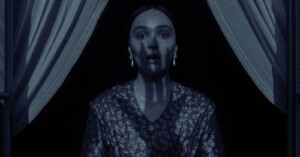 A woman with a surprised expression stands in front of a window with curtains. The image is captured in a dim, blue-toned light, highlighting the details of her patterned clothing and earrings.