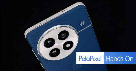 Close-up of a smartphone's rear camera module with three lenses on a sleek, dark blue back. The phone has a metallic frame and a small logo. Text overlay says "PetaPixel Hands-On.