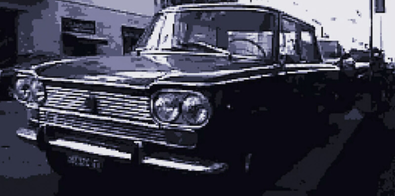 A vintage car with a classic design is parked on a city street. Its gleaming front grille and round headlights are prominent, with buildings in the background. The image has a blue-tinted filter, enhancing the retro appearance.