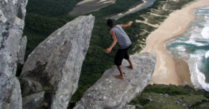 A person in a sleeveless shirt, cap, and shorts balances on a large rock protruding from a cliff. Below is a view of a sandy beach, ocean waves, and dense green forest. The scene conveys a sense of adventure and risk.