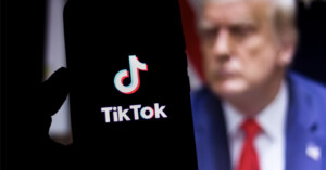 A smartphone displays the TikTok logo on its screen. In the blurred background, a person in a suit with a red tie is visible.