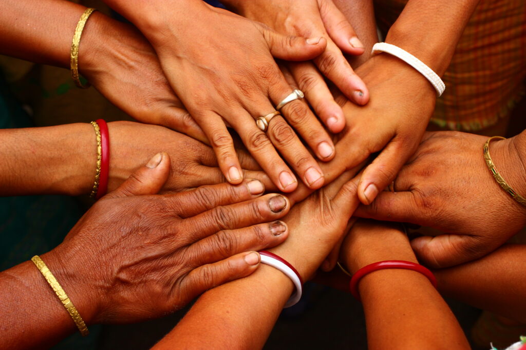 A group of hands of different skin tones and ages coming together in a circle, with some wearing bracelets and rings.