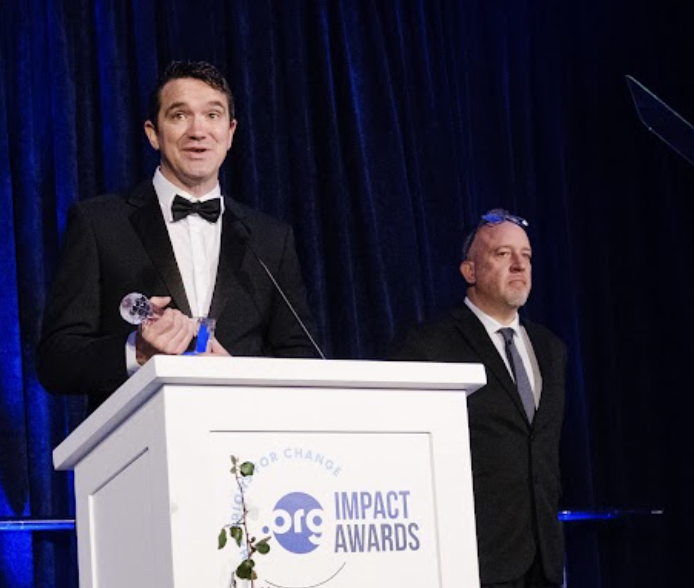 Two men in formal attire stand on stage behind a podium with a logo reading '.ORG Impact Awards.' One man speaks into a microphone while holding a small award.