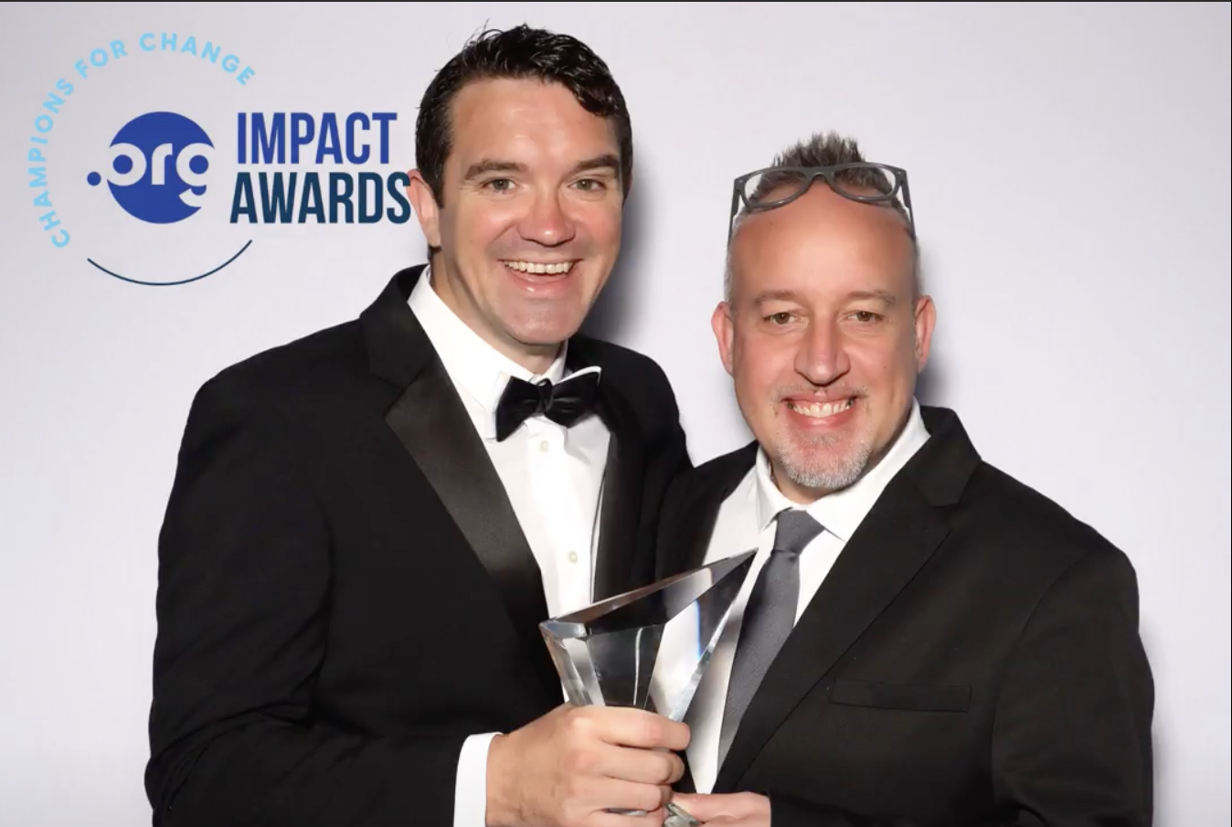 Two men in black suits holding a glass award, with a ".org Impact Awards" logo in the background.