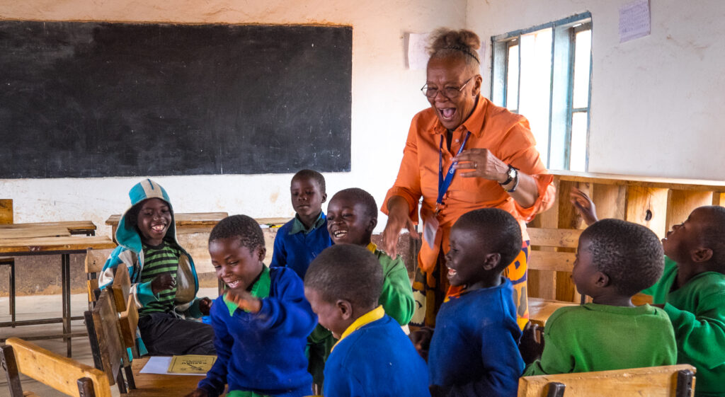 A woman and a group of children in brightly colored uniforms are smiling and laughing in a classroom with a chalkboard in the background.